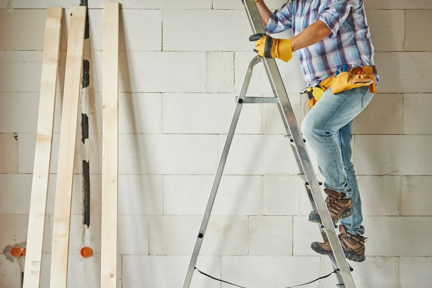 A construction worker in jeans and a plaid shirt climbing a metal ladder inside a partially built room, with wooden beams leaning against a white brick wall, representing home renovation or remodeling work.