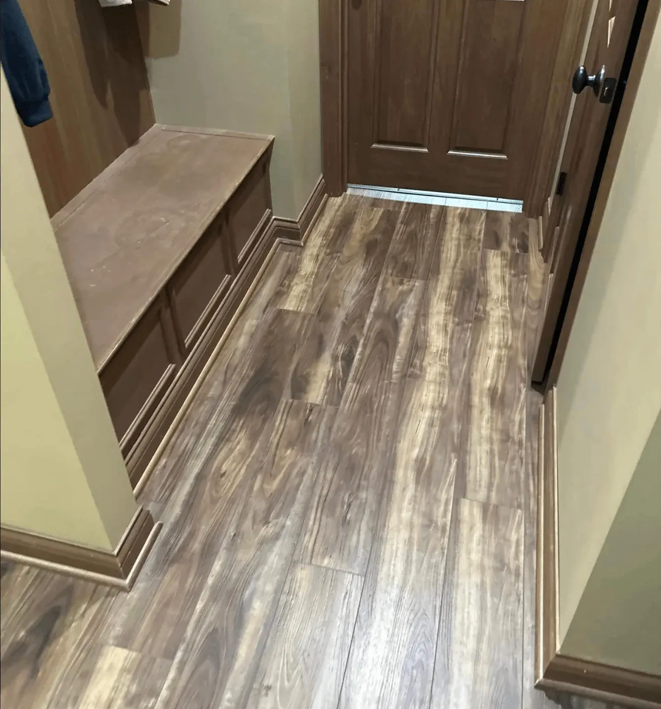 A newly installed wood-look laminate floor in a hallway with brown trim and built-in bench seating, showcasing a warm and rustic home interior design.