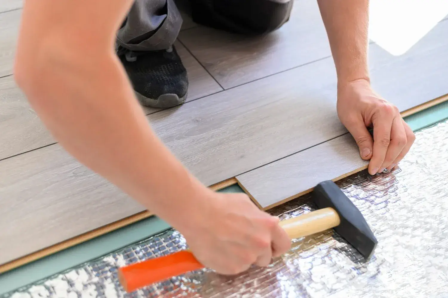 A close-up of a flooring professional installing light gray laminate planks over an underlayment, using a rubber mallet to ensure a snug fit, demonstrating precision and craftsmanship in home improvement.